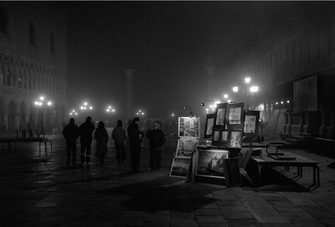 A black and white image of people and an art stall in Venice at night.