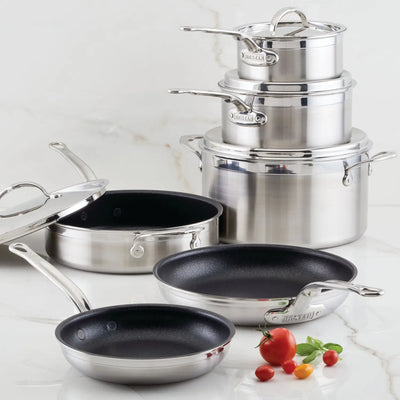 A ProBond® Forged™ Professional Clad Stainless Steel TITUM® Nonstick Ultimate 10-Piece Cookware Set, including saucepans, stockpot with lids, and frying pans, is arranged on a white marble surface with cherry tomatoes and herbs in front.