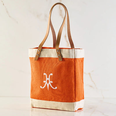 The Hestan Market Tote, Citra Orange by Accessories, is a masterful blend of orange and beige with a resilient design. It stands upright against a marble background, featuring brown leather straps and a white decorative symbol, embodying durable luxury.