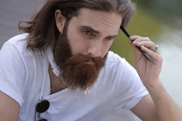 Homem de pele clara, cabelos médios e camiseta branca, na gola da qual está pendurado óculos escuros, encosta caneta na testa, pensativo. Ele usa uma barba estilo Bandholz, castanha com tons ruivos.