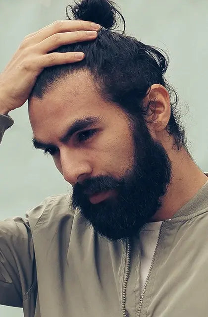 Em artigo sobre o melhor modelo de barba para cada formato de rosto, foto traz homem com barba longa e escura, cabelos escuros e médios em coque samurai, latino, usando agasalho bege e com a mão esquerda na cabeça. 