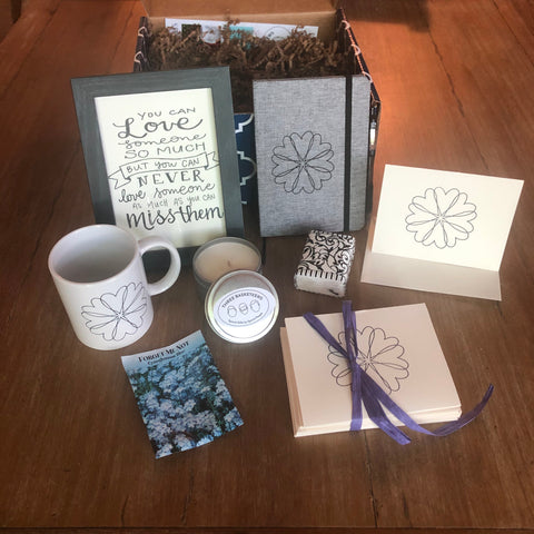 Contents of a Grief Care Basket with a mug, calligraphy, journal, note cards, a candle and Forget-me-not seed packet
