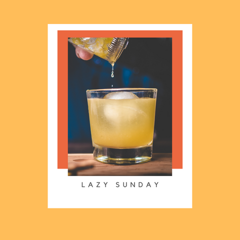 Yellow hued cocktail in a rocks glass with ice against a navy backdrop. Cocktail name is Lazy Sunday.