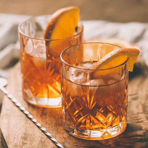 two whiskey cocktails on the rocks with orange slices on a cutting board