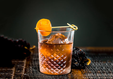 Sophisticated looking cocktail in an old-fashioned glass that has a geometric pattern on it. One large format ice cube and an orange peel on a cocktail pick artfully placed on top. A scattering of real dried ancho chiles in the background on the table.