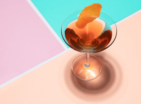 A dark-colored cocktail in a martini glass with an orange twist, against a geometric pastel background