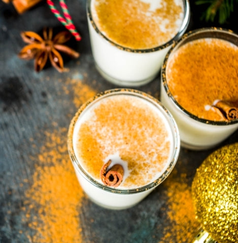 An aerial view of three glasses of frothy eggnog cocktails, with nutmeg sprinkled on top and cinnamon sticks in each. A holiday-themed backdrop in colors of rich brown and gold.