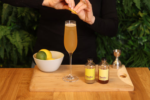 Honey colored cocktail in a champagne coupe, You see the hands of woman garnishing the cocktail with a lemon peel curl.