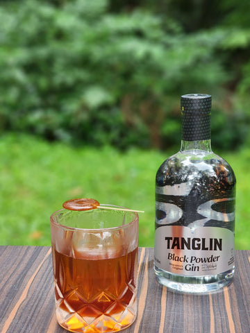 Foreground is a warm, dark orange colored cocktail in a rocks glass with what looks like a lollipop on top.  Background is a bottle of Tanglin Blackpowder Gin.  Both are resting on a wood table with grass and greenery behind.