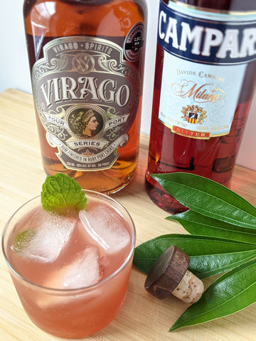 shot taken from above with liquor bottles Campari and Virago Four Port Rum in the background, a pink cocktail garnished with a mint leaf in the foreground.