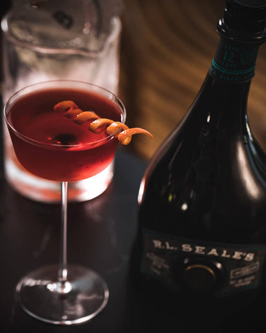 A dark red cocktail with orange twist in a sophisticated glass with a bottle of R.L. Seale's 12 Year rum next to it