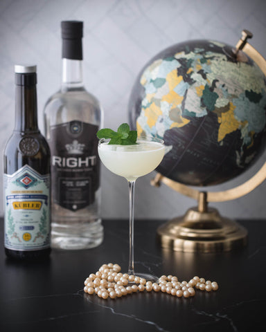 An image of a cocktail in a thin stemmed glass with absinthe and gin bottles and a globe and pearls as the backdrop