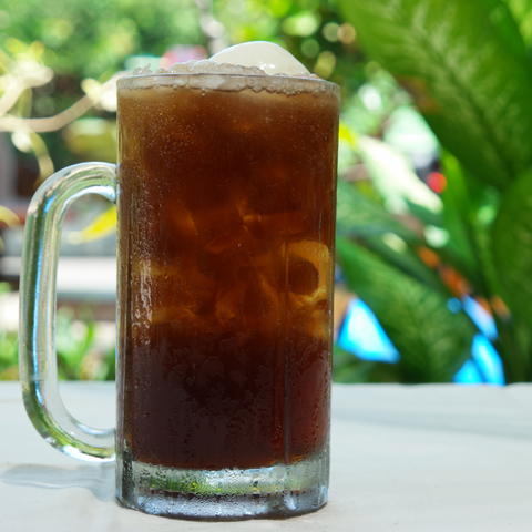 Frosty beer mug in a tropic setting filled with what looks like dark liquid, ice, and ice cream on top.