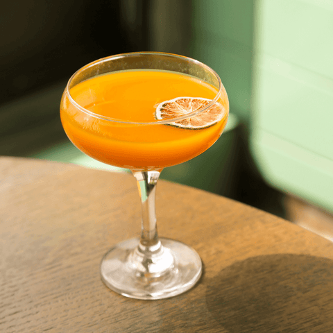 A beautiful orange-hued cocktail in a coupe glass with a lime wheel floating on top and a pistachio green backdrop