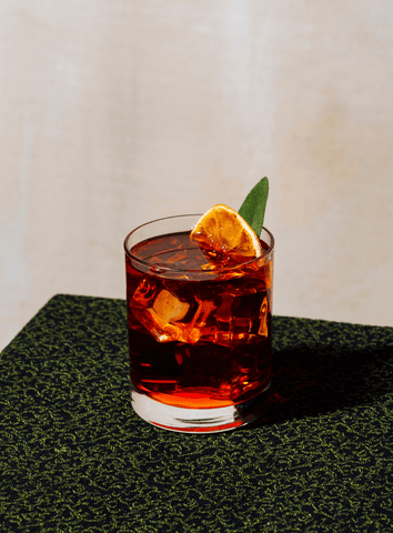 The Banana Boulevardier is a rich red cocktail in a rocks glass on a neutral backdrop with an orange garnish.
