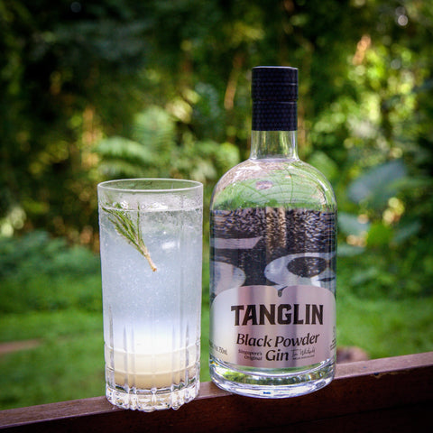 A cocktail in a Tom Collins glass on the back deck with a bottle of Tanglin Black Powder Gin beside it. Refreshing, cool, a sprig of rosemary to garnish.