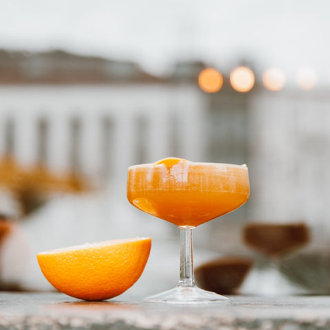 an eye level view of a peach colored cocktail in a coupe in the foreground, with half an orange sitting to its left. Out of focus architectural shapes in the background. 