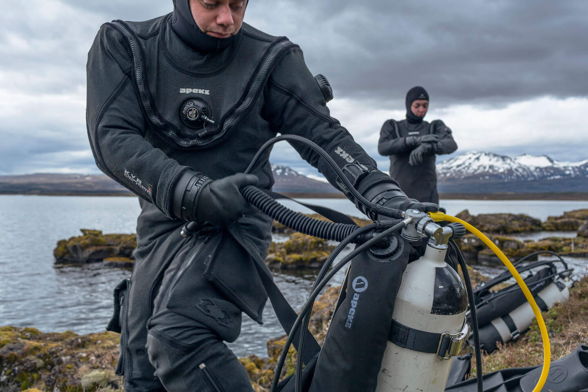 Man Wearing Apeks Drysuit putting on a scuba diving rig by the sea 