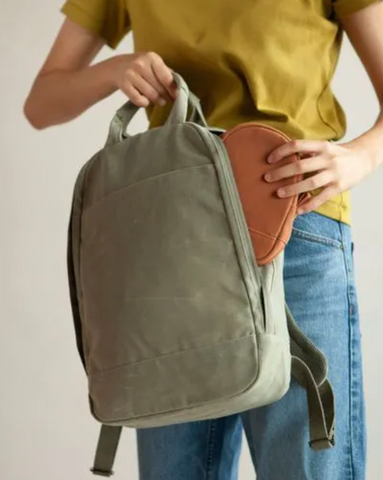 a picture of the the sustainable Day Owl laptop backpack in olive green. A woman is putting a maple brown pouch into the bag while holding the backpack from the top handles