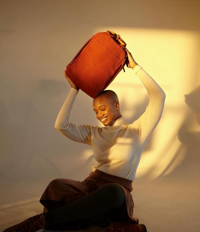 a black women with short hair wearting a white sweater and love velvet dress holds a Day Owl laptop backpack over her head while sitting on the floor.