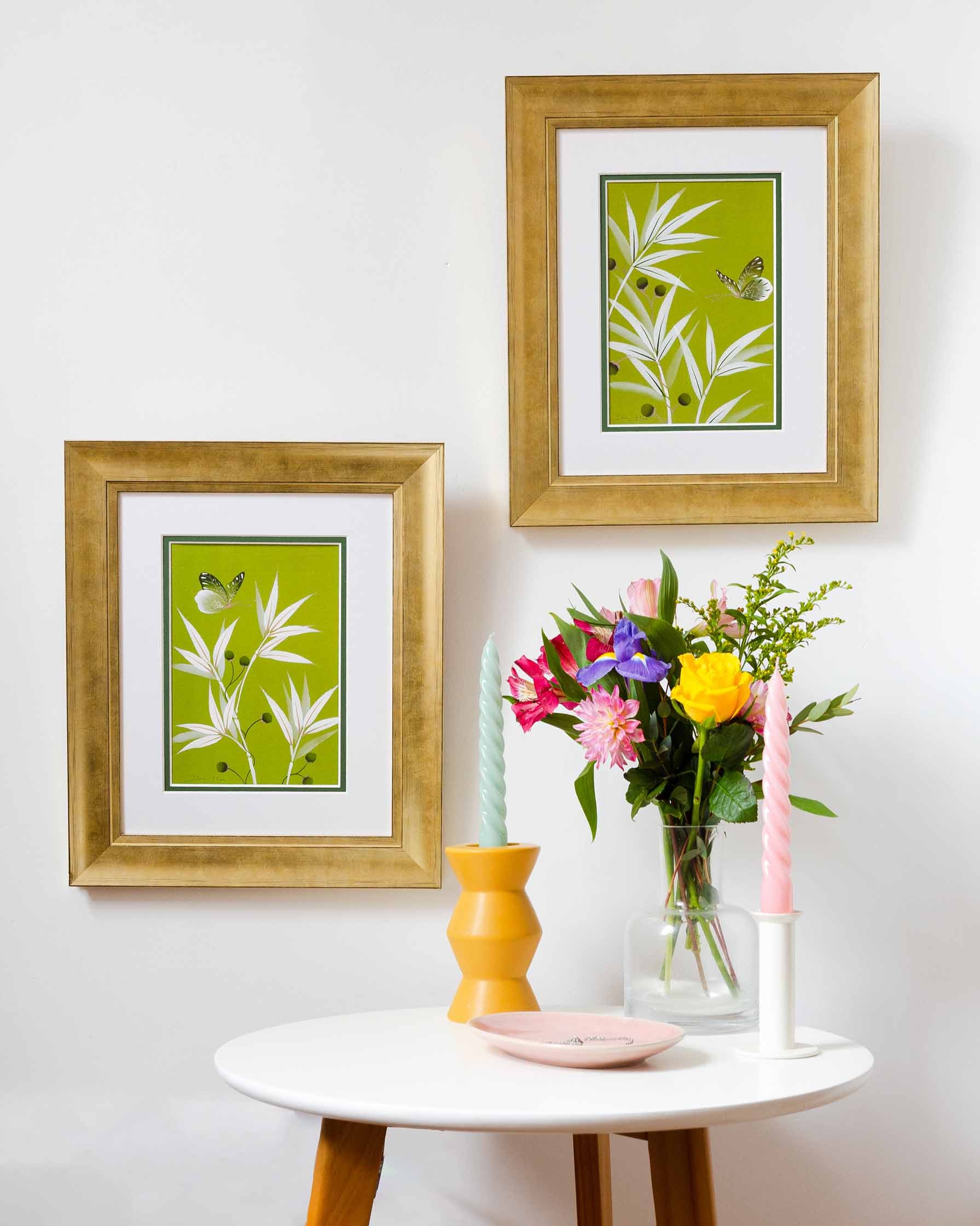 Diane Hill's prints 'Clarissa & Joanna' in gold frames on a white wall with a table containing flowers and candles beneath it