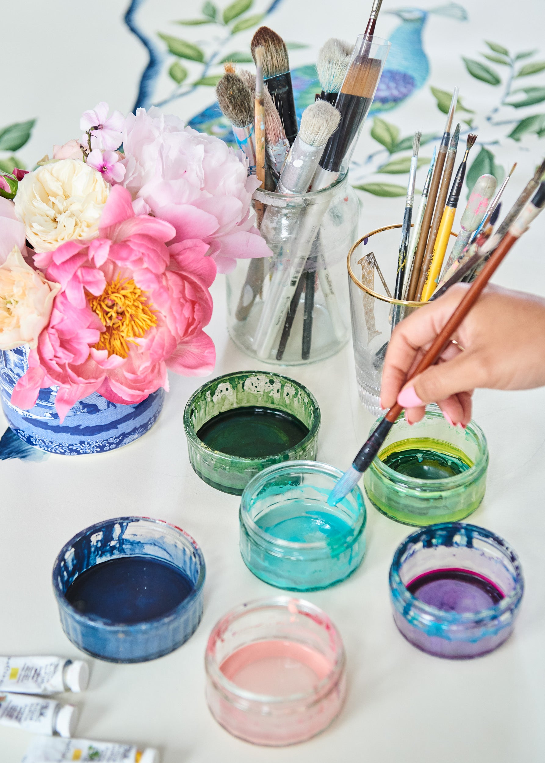 diane hill dipping her paintbrush into a pot of watercolour paint surround by other pots of paint next to a vase of flowers