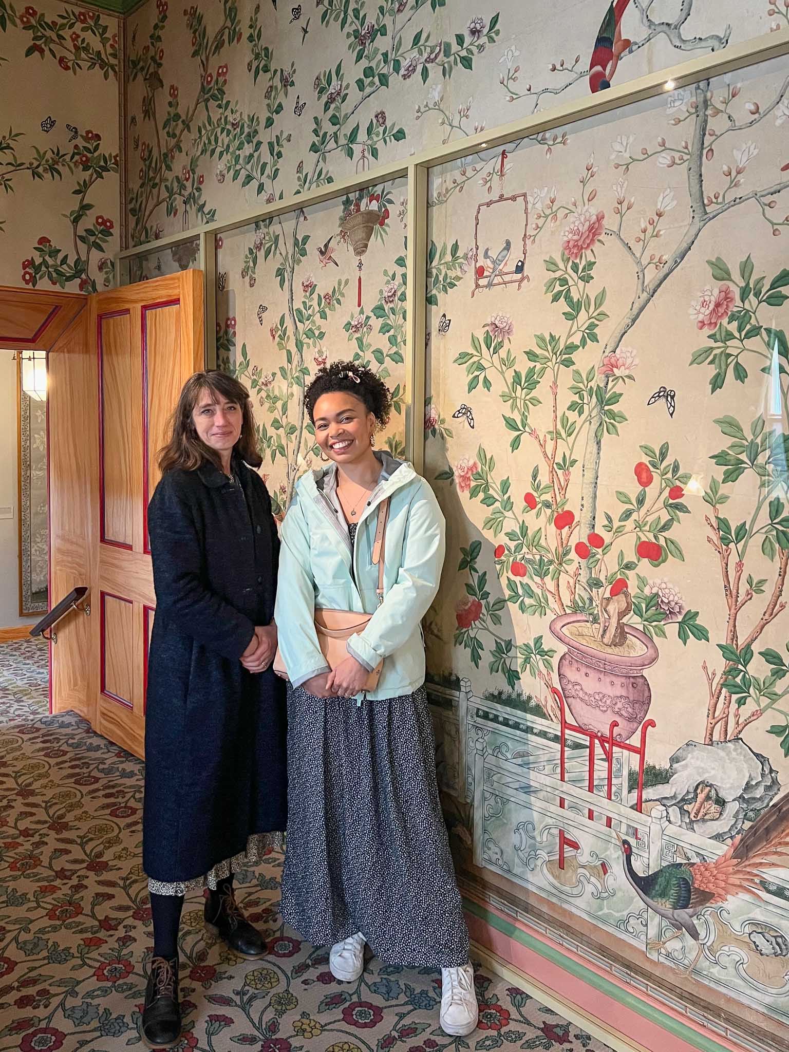 Artist Diane Hill and art historian Dr. Alexandra Loske in front of colourful chinoiserie wallpaper inside Brighton Pavilion