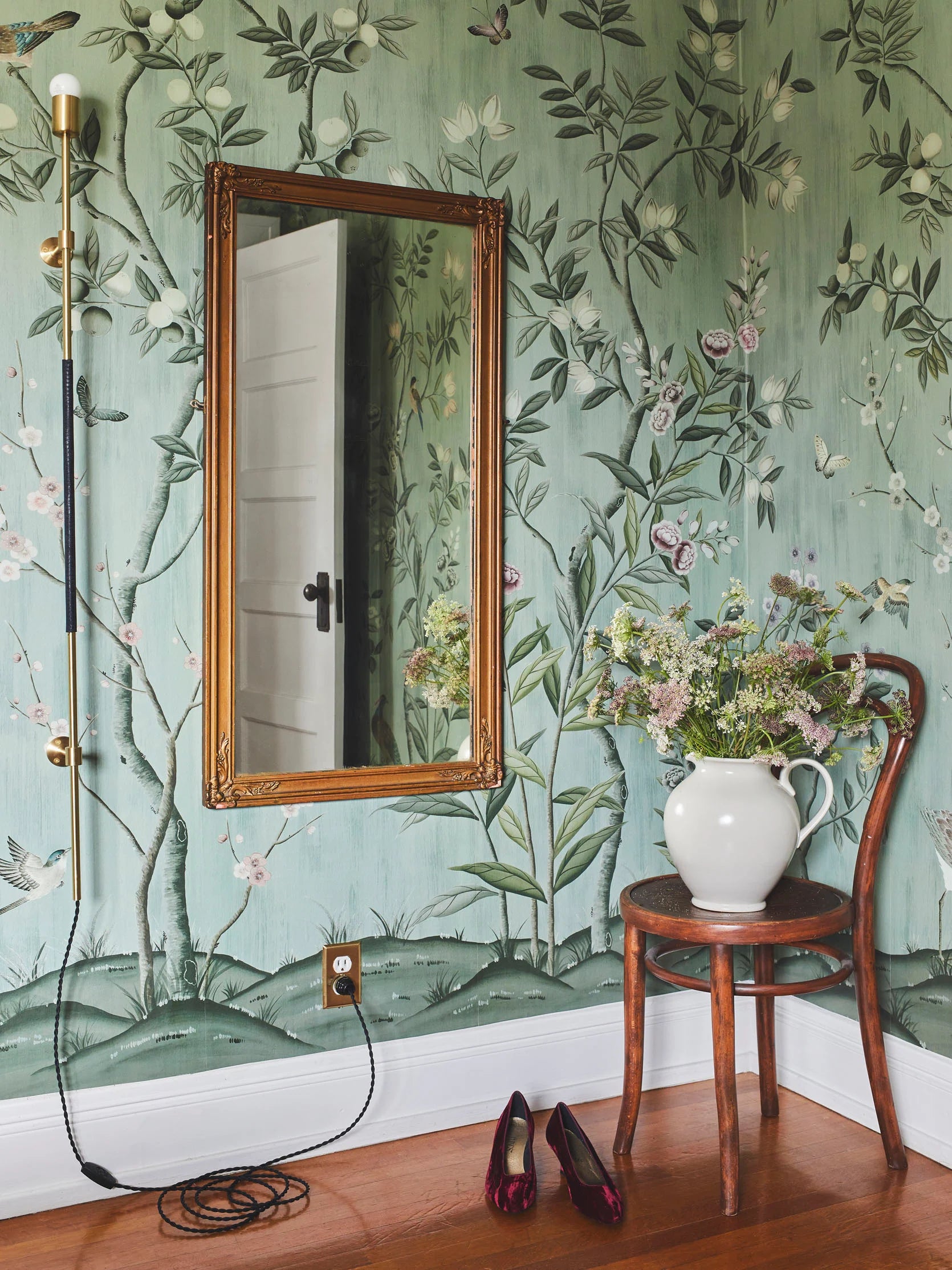 bedroom featuring Diane Hill's 'chinoiserie chic, jade' wallpaper for rebel walls, antique mirror, vintage style tall lamp, brown antique looking chair with a vase of flowers on top and a pair of shoes