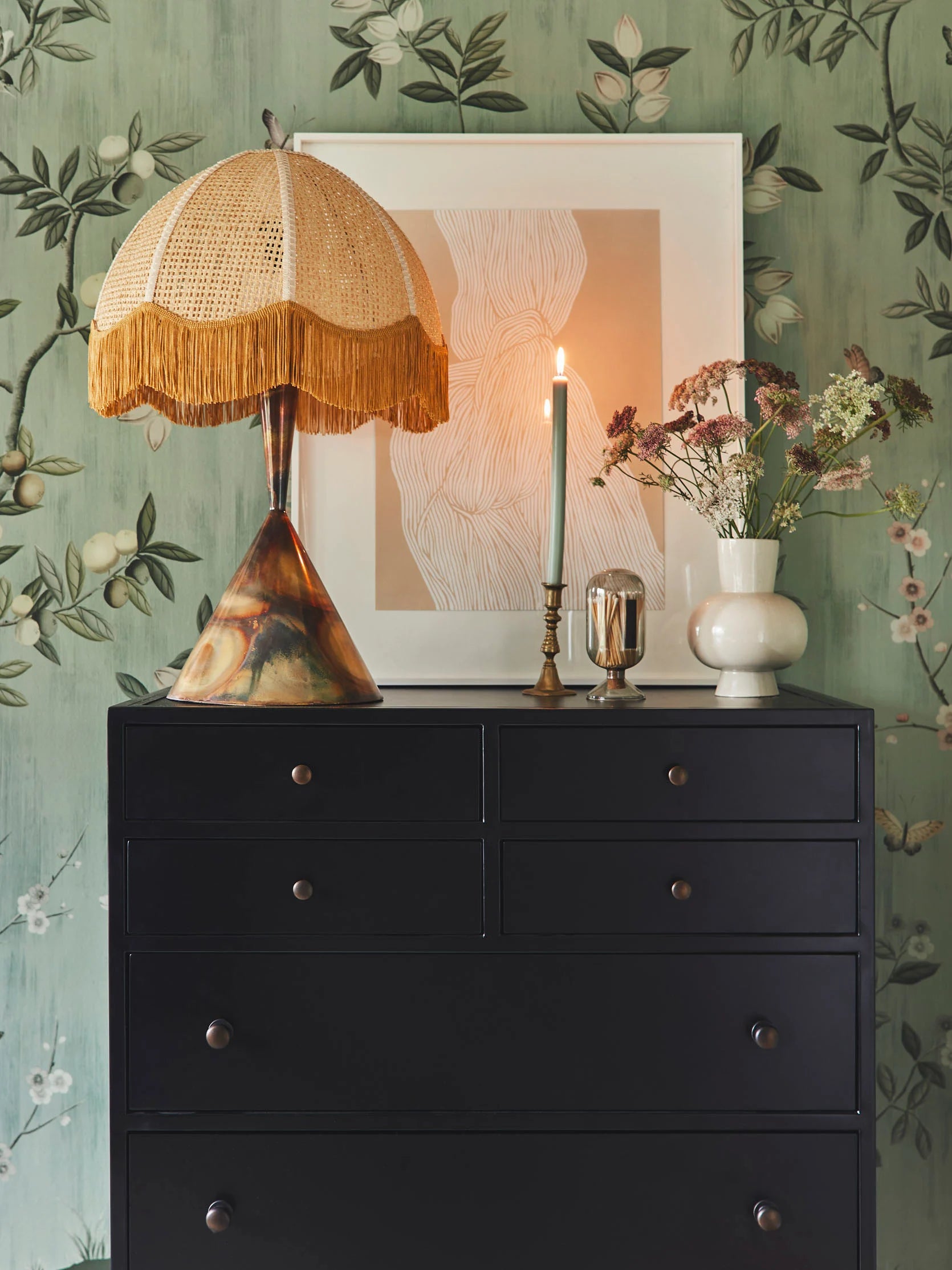 Maximalist home decor. Set of dark wood draws with a lamp, candle, ornaments, and art print on top in front of green chinoiserie wallpaper.