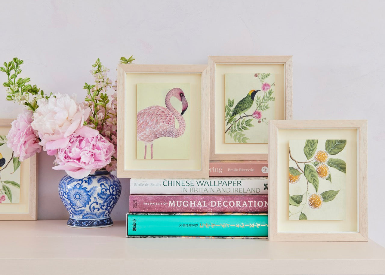 three of Diane Hill's mini botanical prints containing images of chinoiserie style birds and leaves in frames on top a desk with books and a vase containing flowers