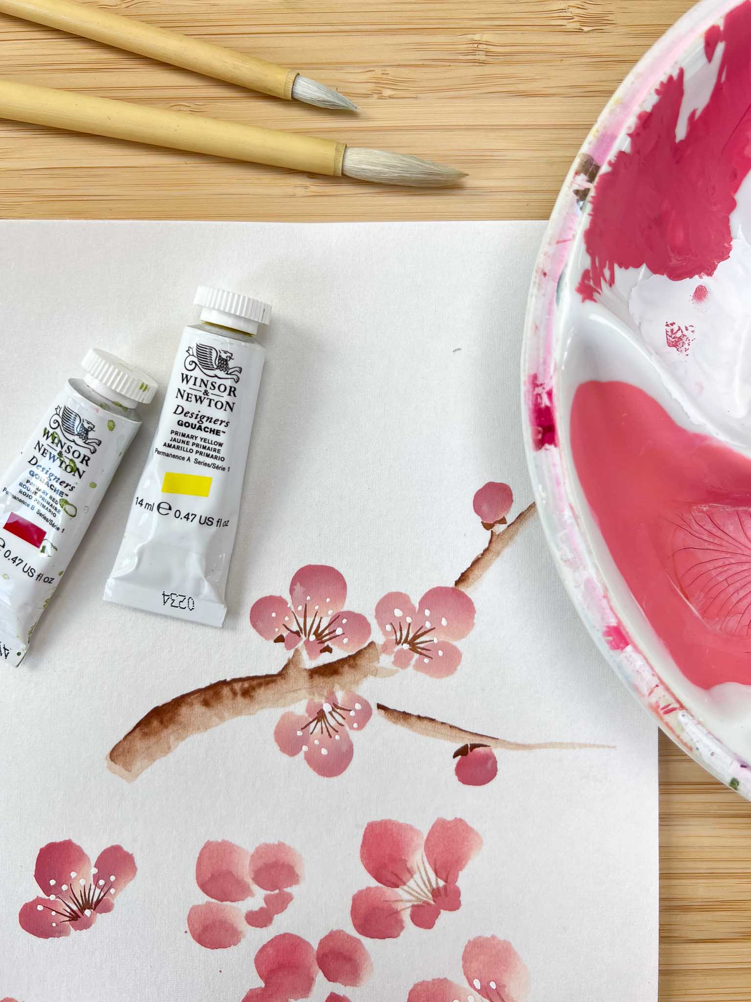 paintbrushes, paints, and a palette next to a painting of pink blossom flowers on silk paper