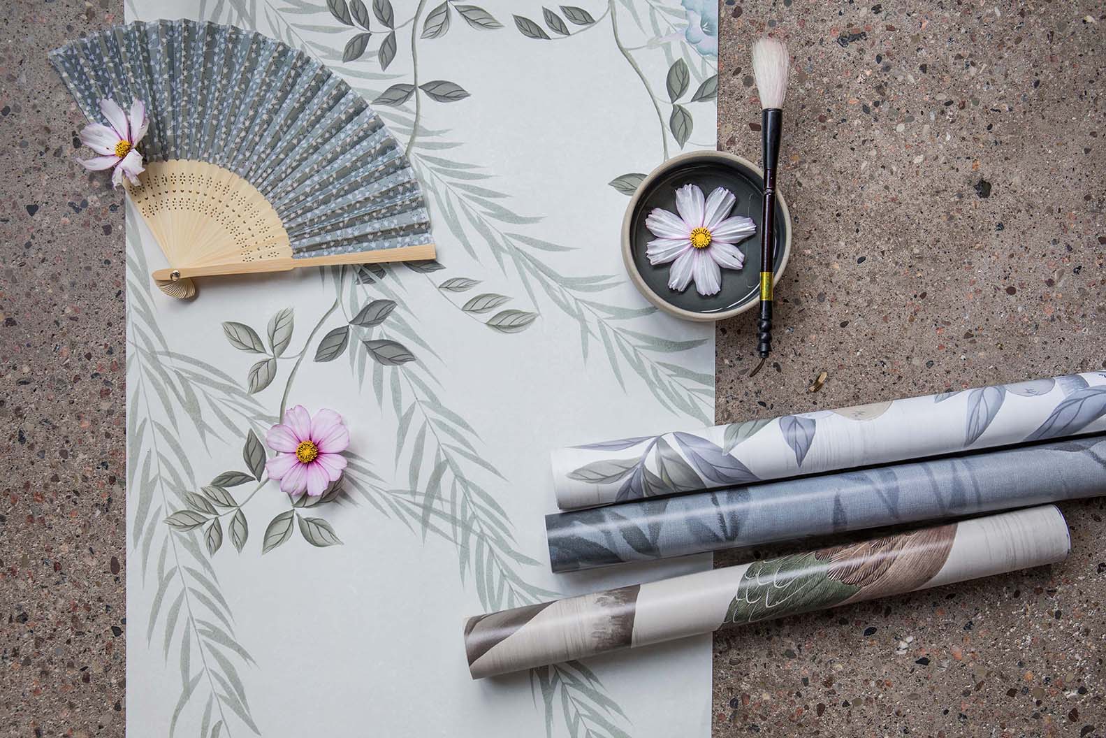 Chinoiserie wallpaper designed by Diane Hill for Rebel Walls' 'La Chinoiserie' collection panel laid flat with an oriental fan, flowers, and other rolls of wallpaper laid on top