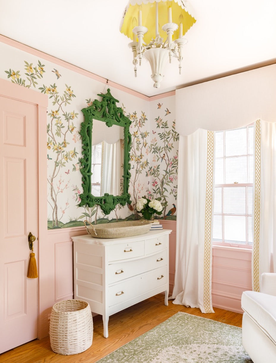 Grandmillenial style nursery room with a green mirror on a wall containing 'Chinoiserie Chic' wallpaper by Rebel Walls designed by Diane Hill. Room contains white vintage furniture and a powder pink door.