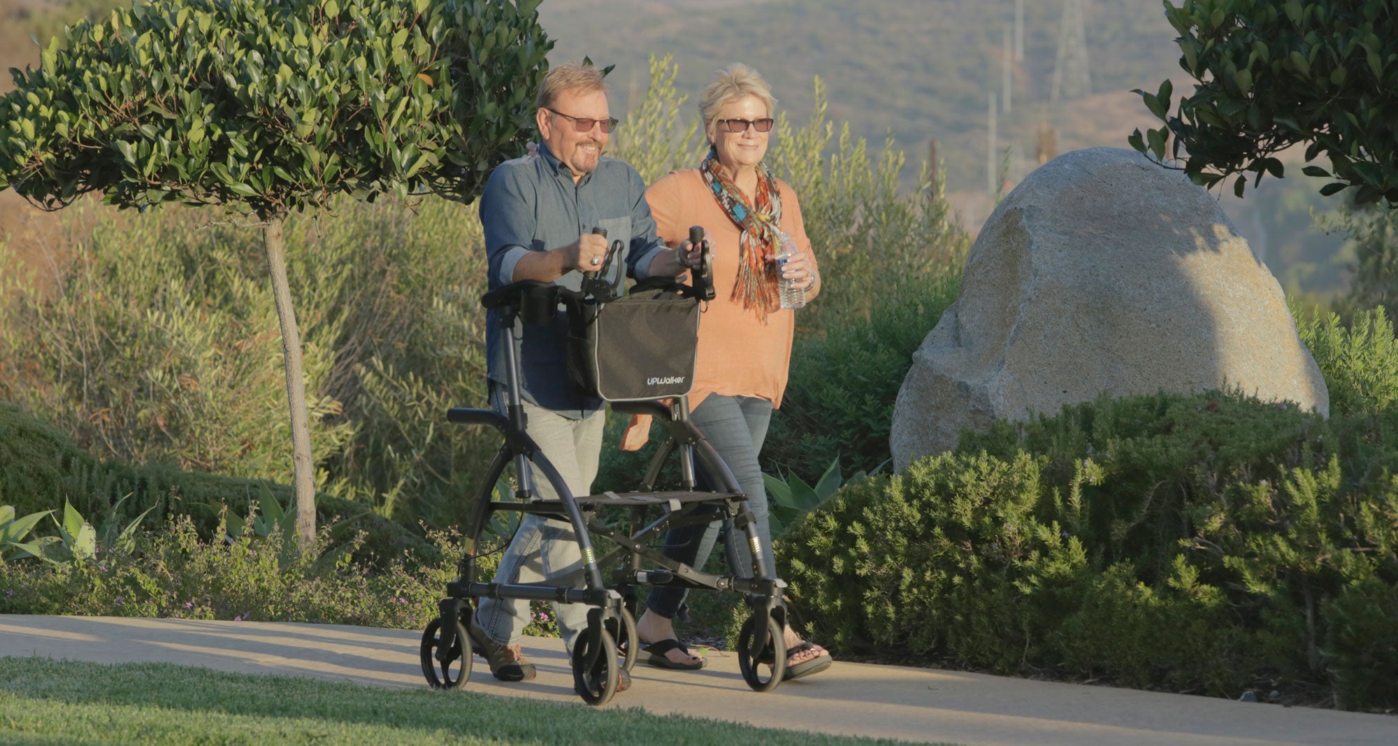 A man smiling as he walks through a sunny park with a woman and using an UPWalker upright walker with a seat from LifeWalker.