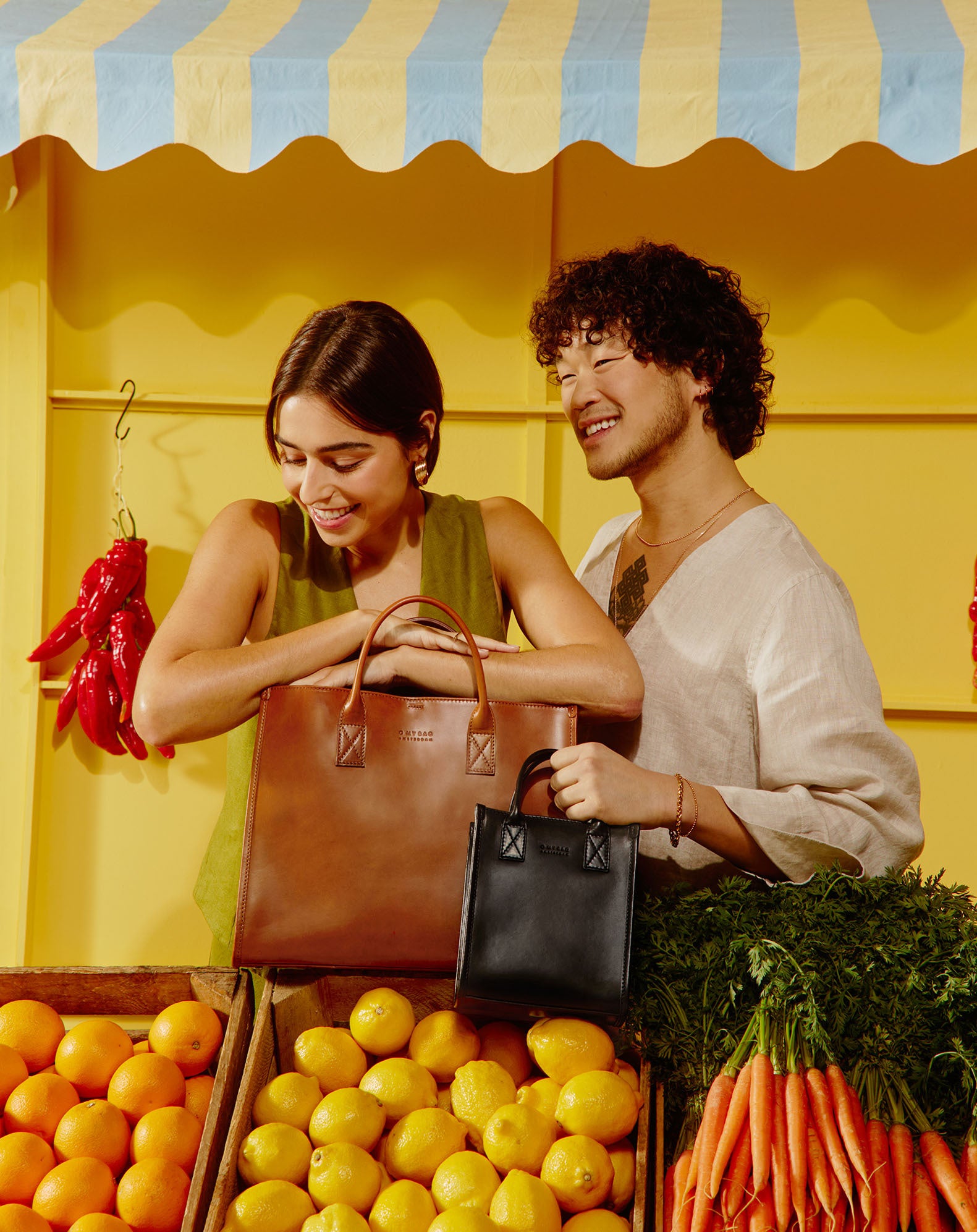 Campaign image featuring fresh fruit and citrus colours, a male and a female model wearing black and brown square tote bags