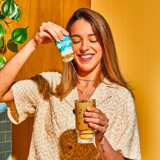 A smiling woman adding a squeeze of Buoy hydration drops to her glass, enhancing her beverage for better hydration and digestive health.