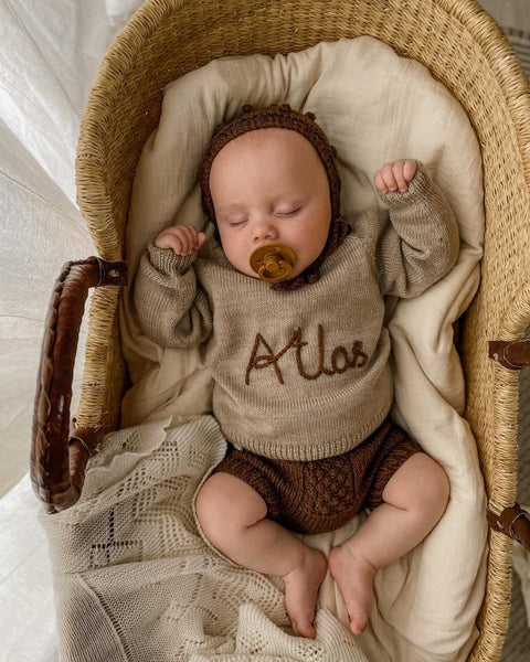 Baby sleeping in our personalized merino wool sweater in the basket. The sweater is matched with knitted shorts and a bonnet.