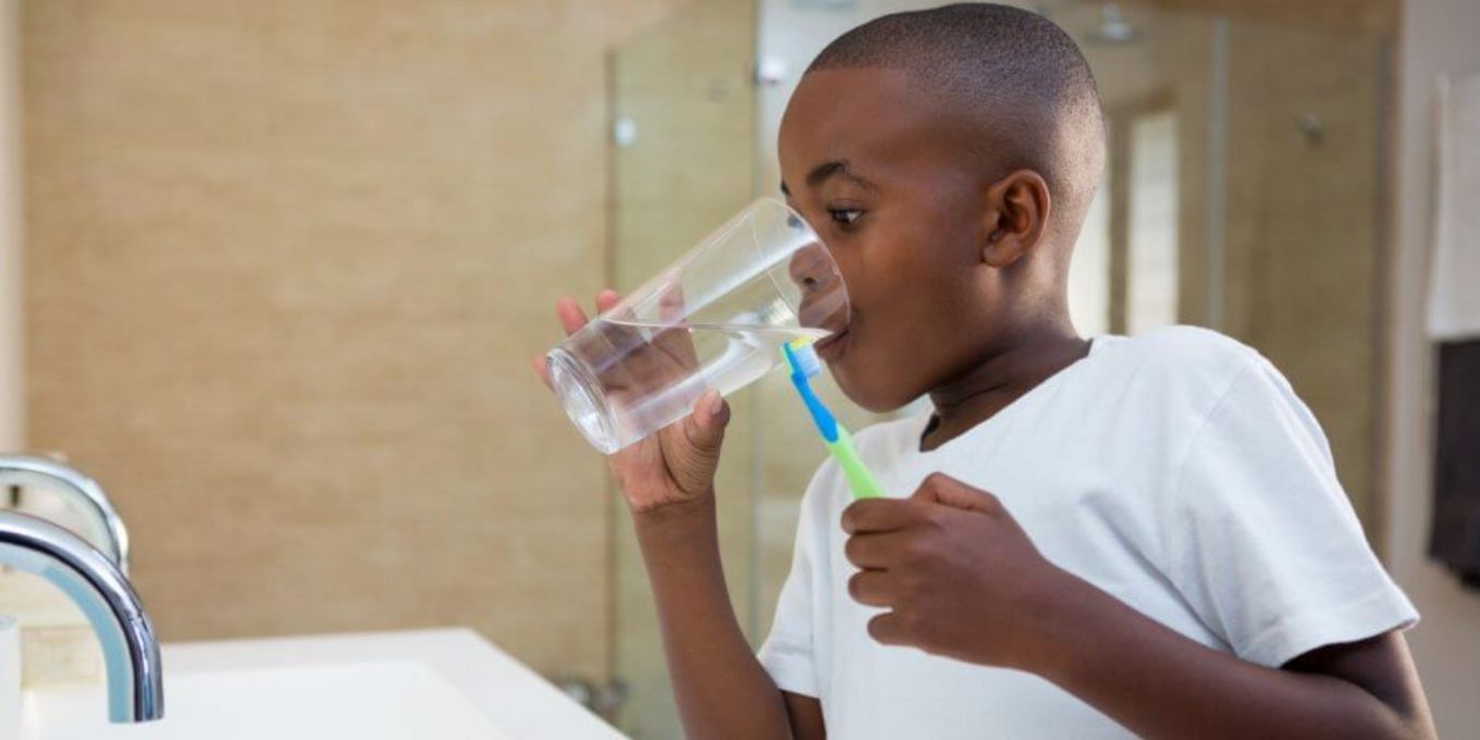 Little boy drinking water from a glass with a toothbrush in his hand