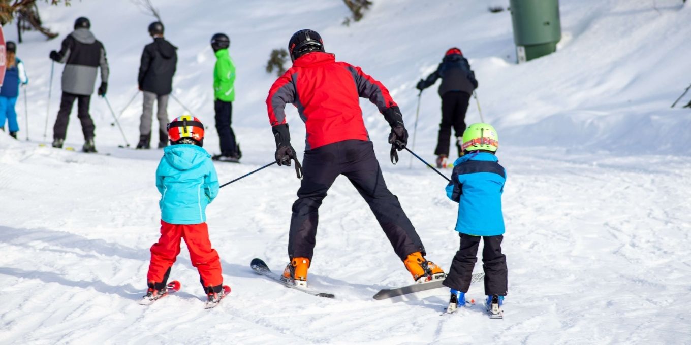 Father skiing down bunny hill with 2 kids.  Each child is holding onto the father's ski poles