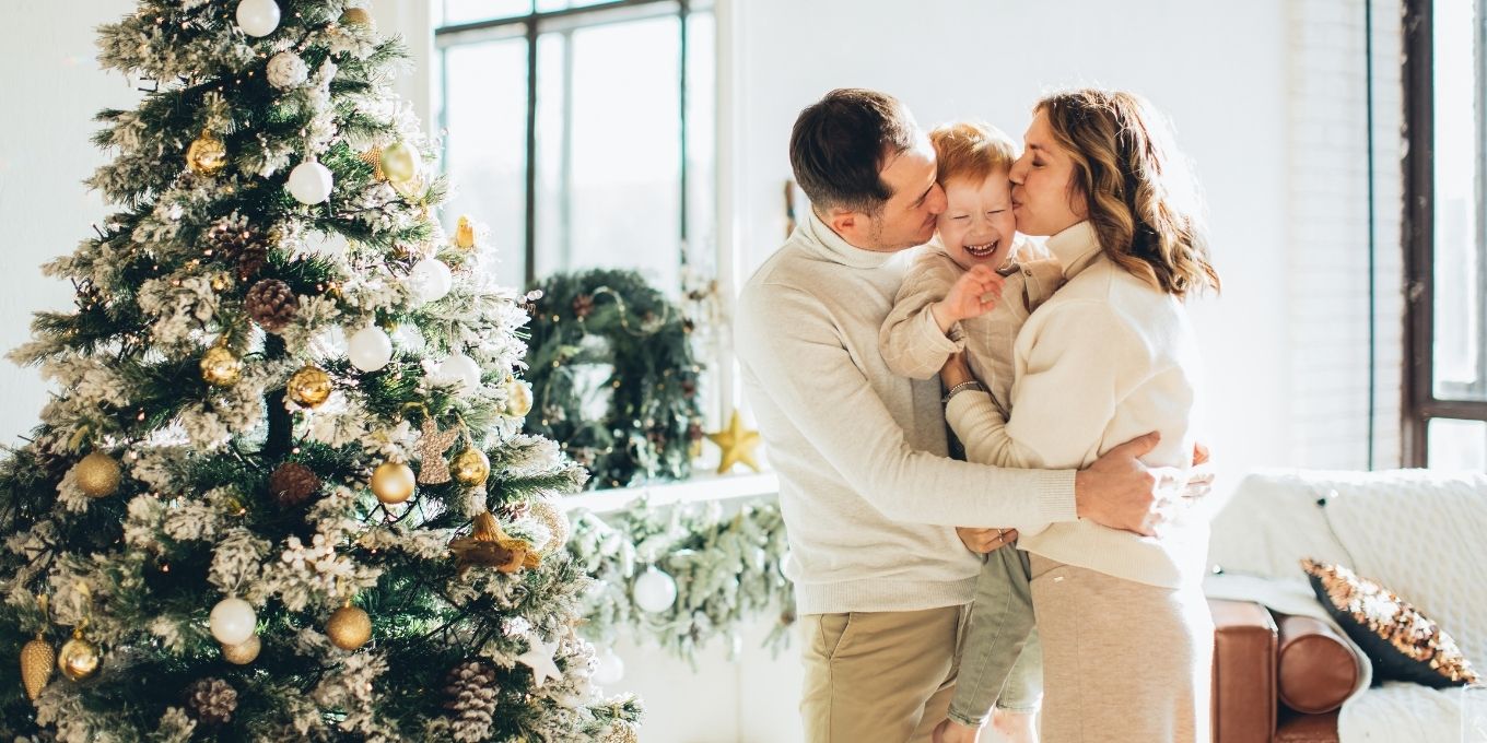 Parents holding son together kissing him on the cheek with Christmas tree in the background