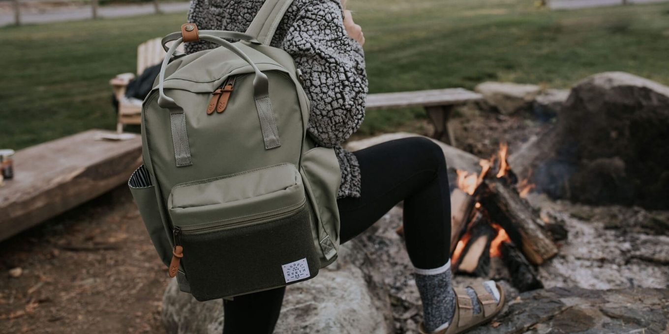 Woman standing in front of a fire pit carrying a Sage Elkin Diaper Bag Backpack on her right shoulder