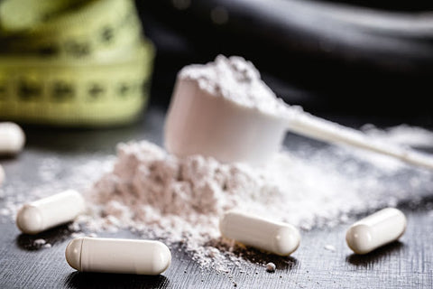 A photo of creatine pills on a table in front of a mound of creatine powder with a measuring cup full of creatine powder next to a rolled up measuring tape.