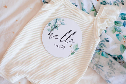 baby milestone card with text written "hello world" sitting on a baby onesie.