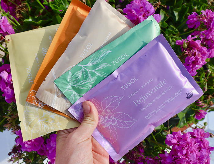 A hand holding colorful TUSOL smoothie mix packets against a backdrop of pink flowers.
