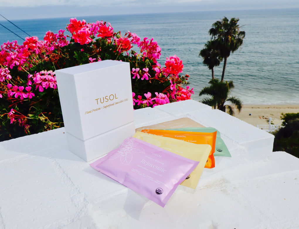 Health smoothie packets and a box from TUSOL on a wall with blooming pink flowers and a beach background.