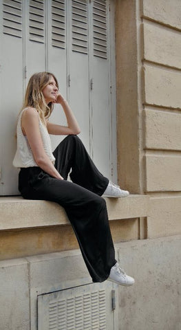 Mannequin qui prend la pose devant une fenêtre avec le pantalon noir et ample "Rio" de la marque française et engagée Nualé