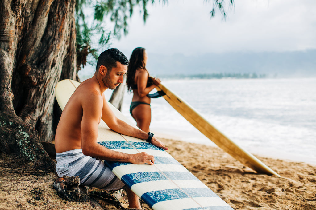 man posing for a photo while sitting on the beach looking down at his blue and white surfboard with a woman looking out into the ocean holder her board behind him