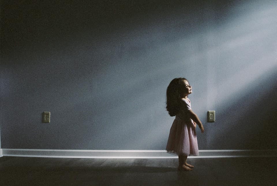 A young girl in a pink dress basks in the sunlight coming in from a window in a real film photo taken on fuji 400h film.