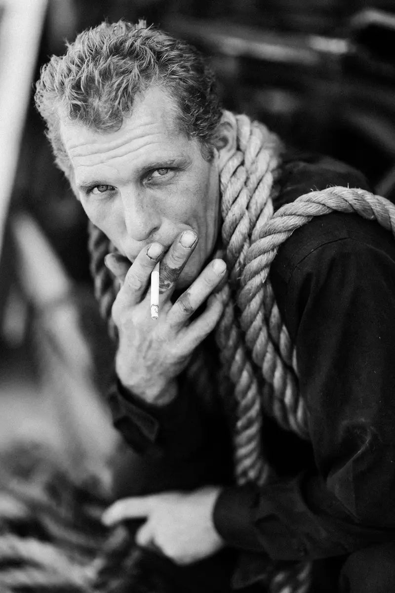 man leaning over smoking a cigarette with ropes wrapped around his shoulders posing for a picture