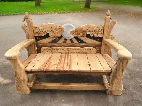 Front view of a handcrafted wooden bench with a detailed carved backrest depicting clouds, a crescent moon, and two perched birds. The backrest is engraved with a heartfelt message dedicated to Gary Bolton